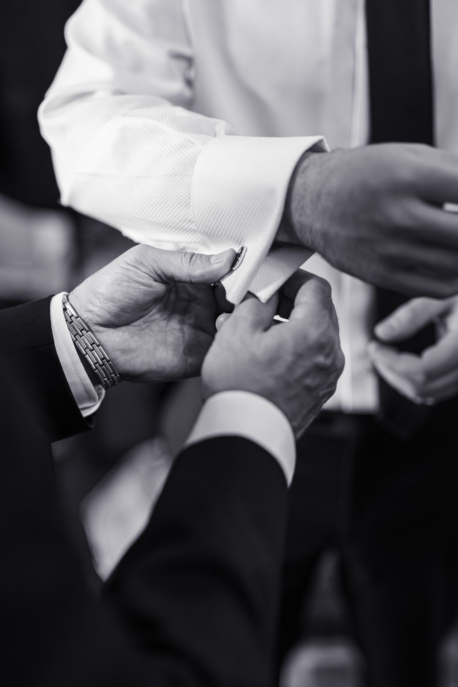 Black and white photo of a man having his cufflinks fastened by another person in formal attire.