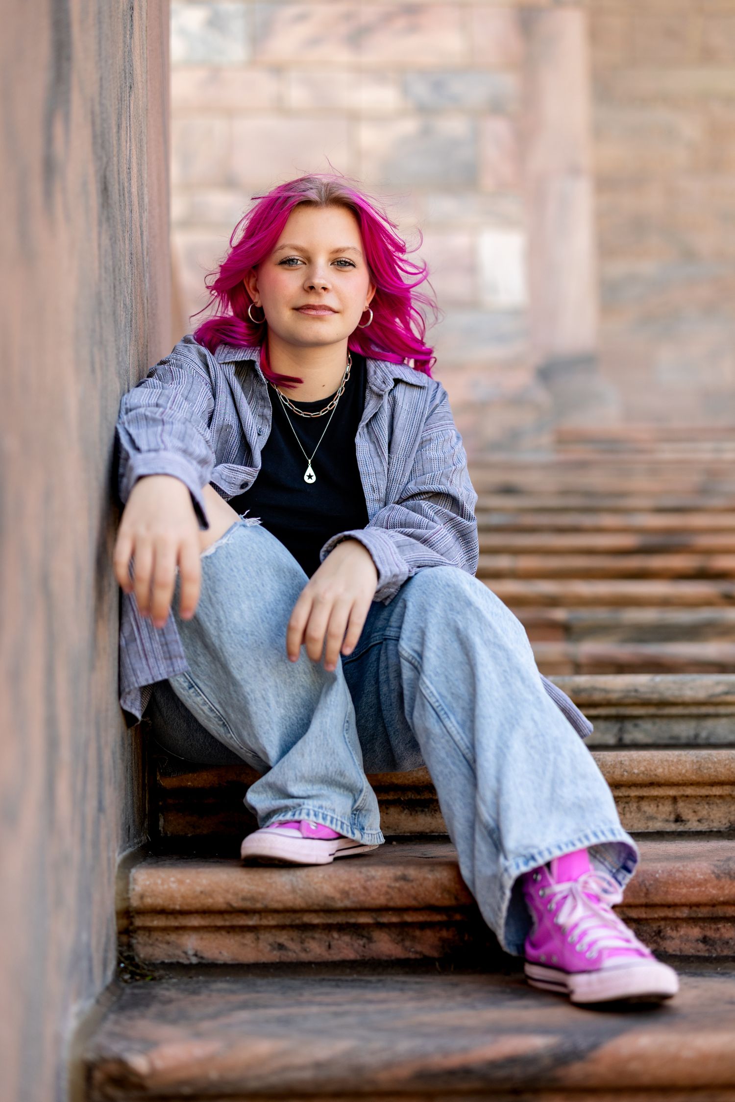 Young woman with pink hair sitting on stone steps wearing jeans, blue shirt, and pink Converse sneakers.