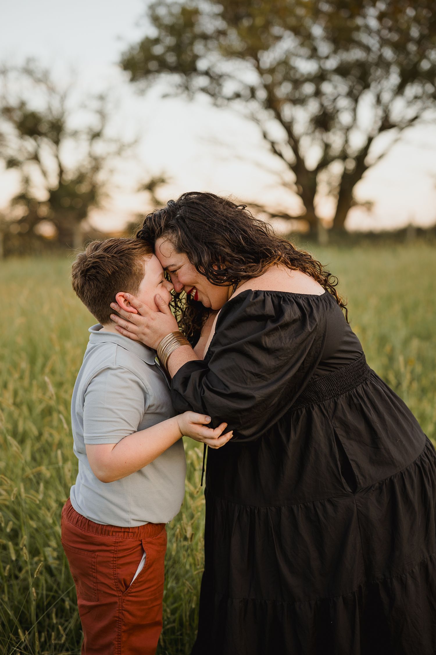 A woman in a black dress shares a tender moment with a young boy in a green field at golden hour.