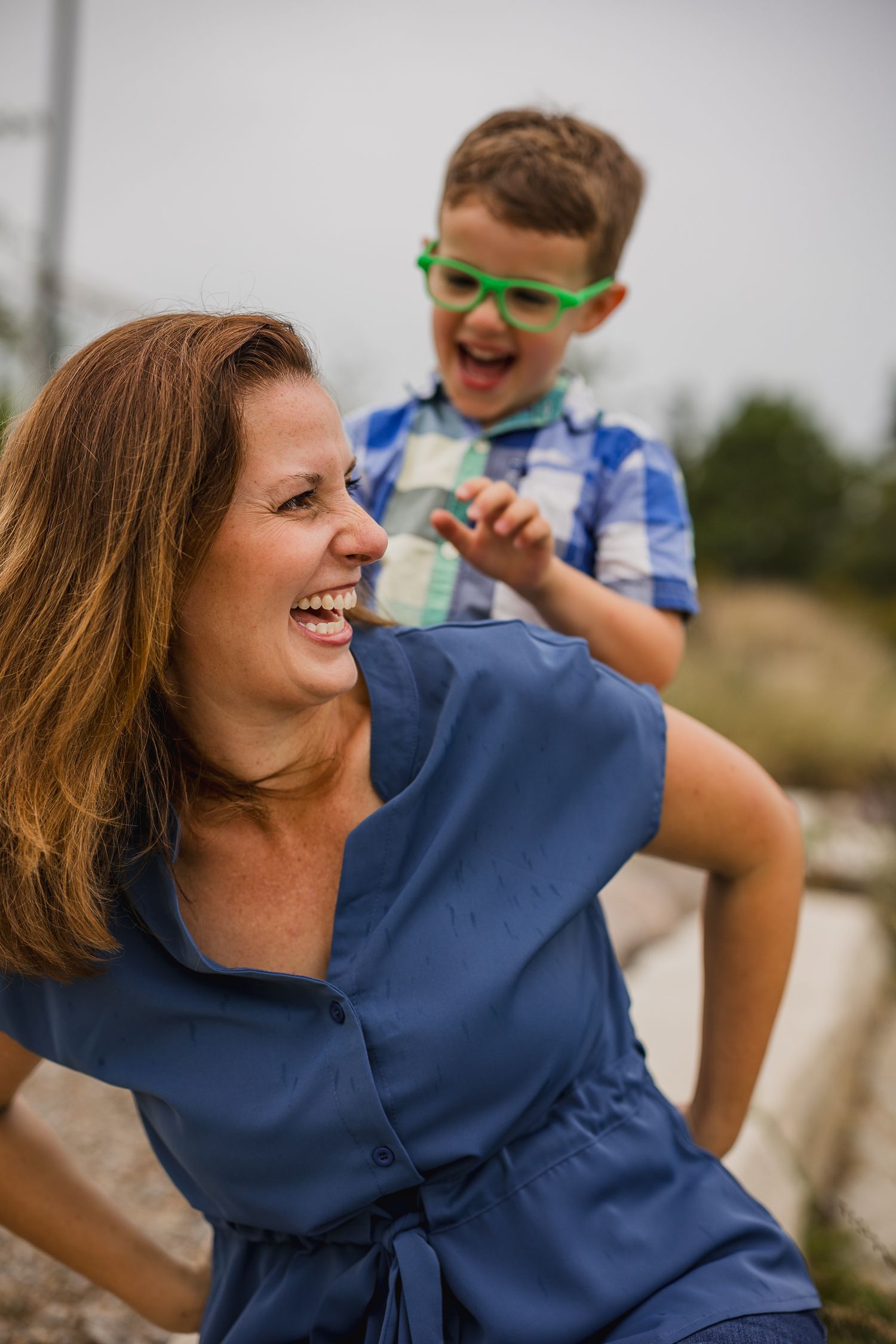A laughing woman in blue dress plays with an excited boy wearing green glasses outdoors.