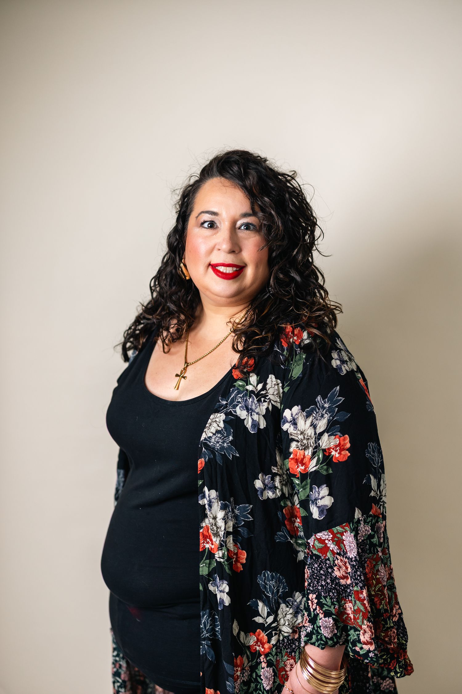 Woman with curly hair wearing floral kimono and black top, smiling confidently against neutral background.