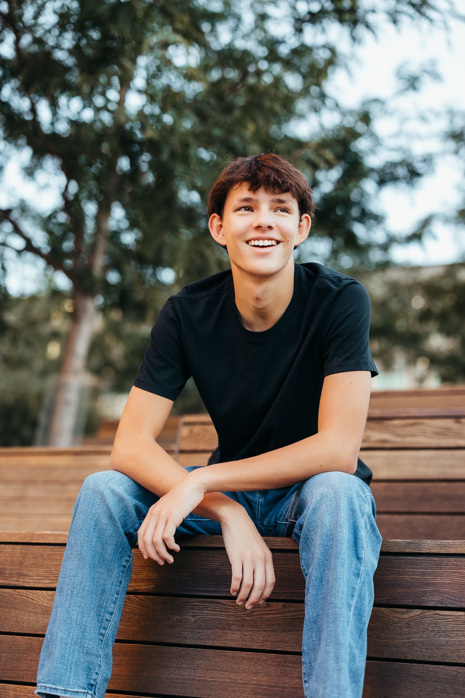 Smiling teenage boy in black t-shirt and jeans sitting on wooden deck outdoors with trees in background.