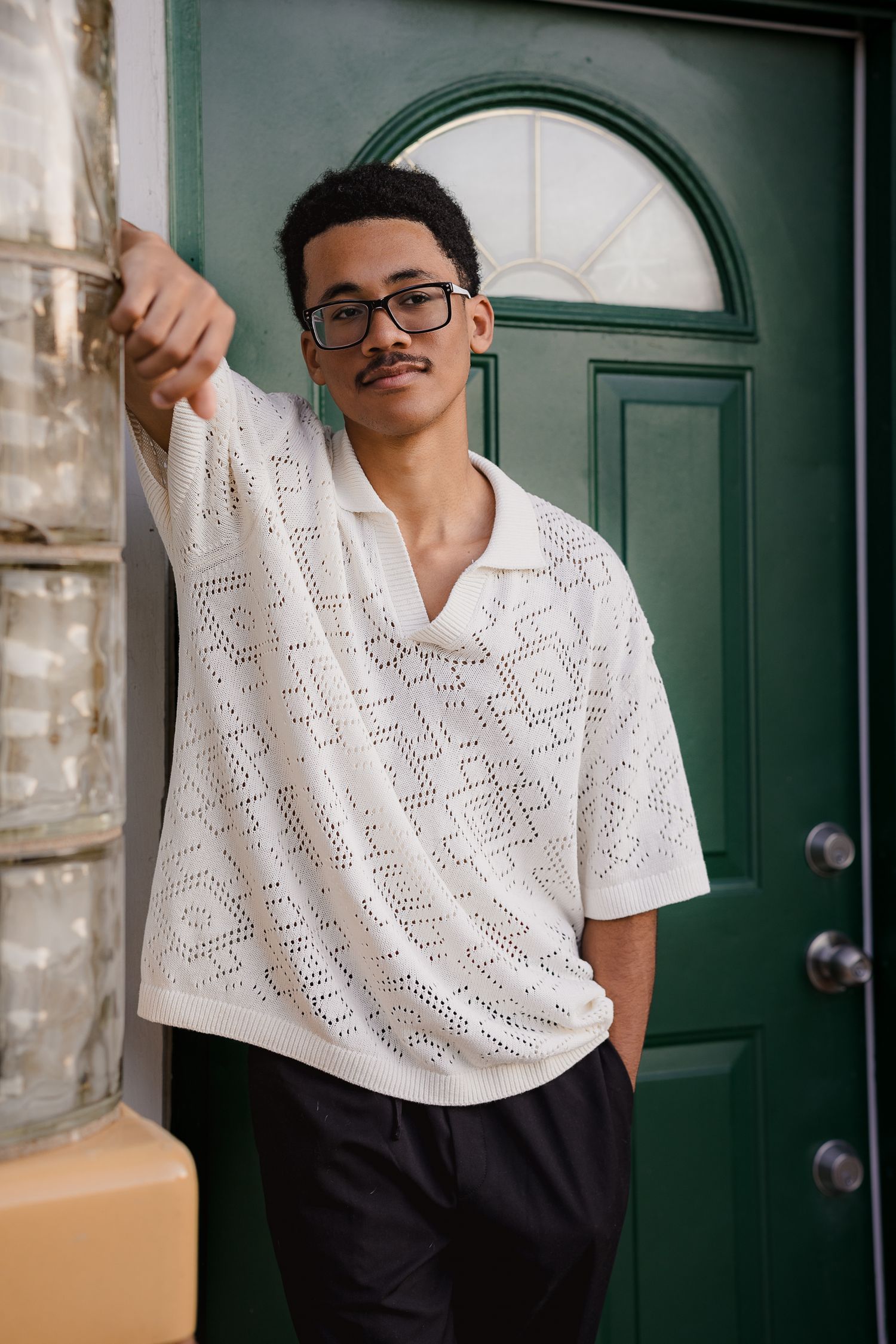 Young man in white crochet polo shirt and glasses leaning against green door in urban setting.