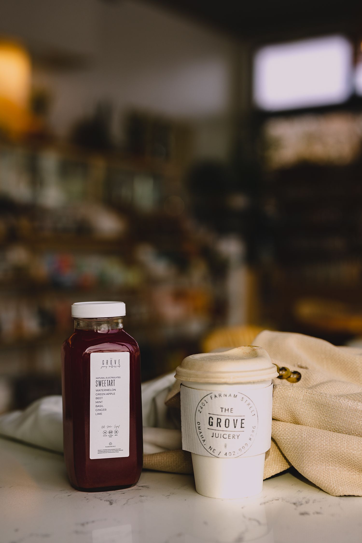 A cold-pressed juice bottle and The Grove Juicery coffee cup on a marble counter in a cozy cafe.