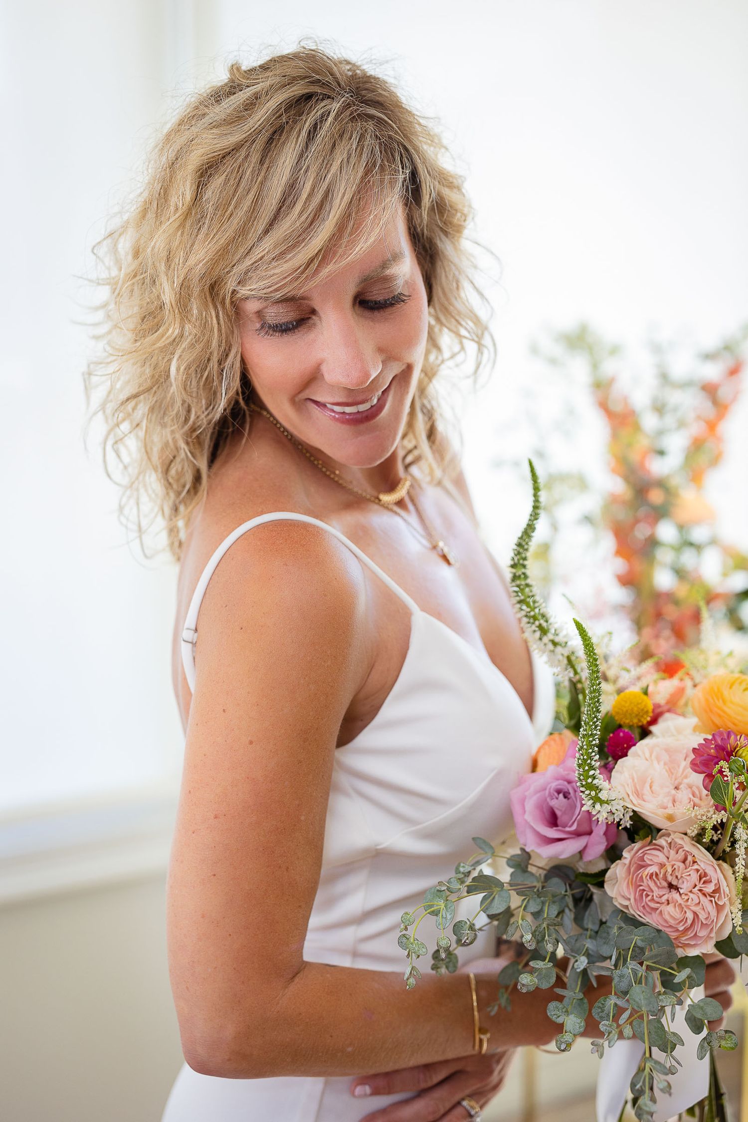 Smiling bride with curly blonde hair in white dress holding vibrant pink and orange wedding bouquet.