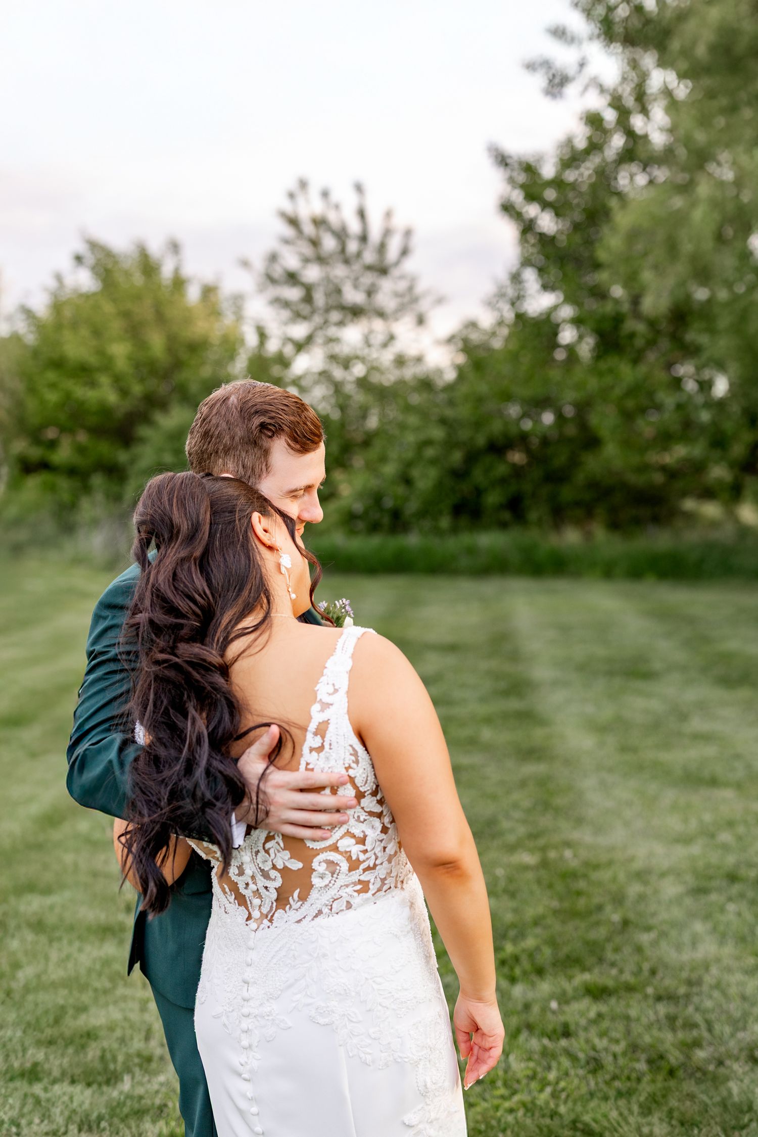 A bride and groom embrace outdoors on a green lawn, the bride in a lace backless gown as the groom kisses her head.