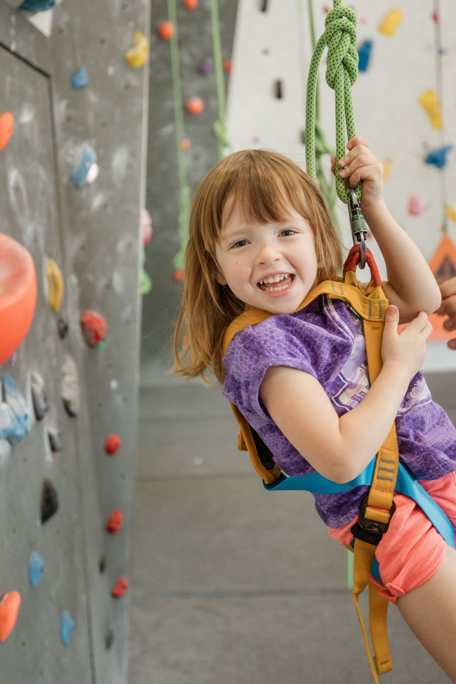 Happy young girl in harness smiling while holding rope at colorful indoor rock climbing wall.