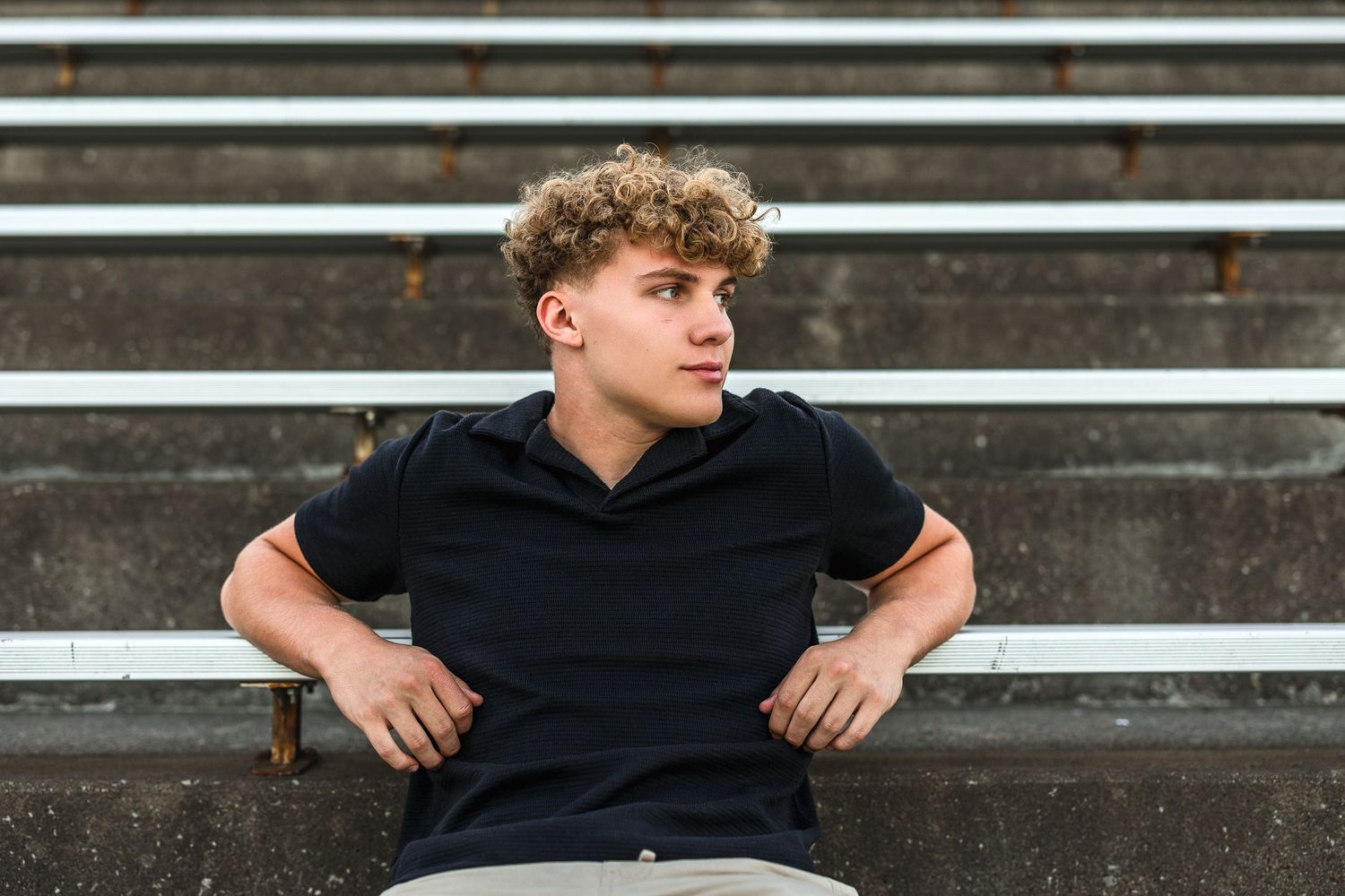Young man with curly hair sitting on bleachers wearing a black polo shirt, looking to the side.