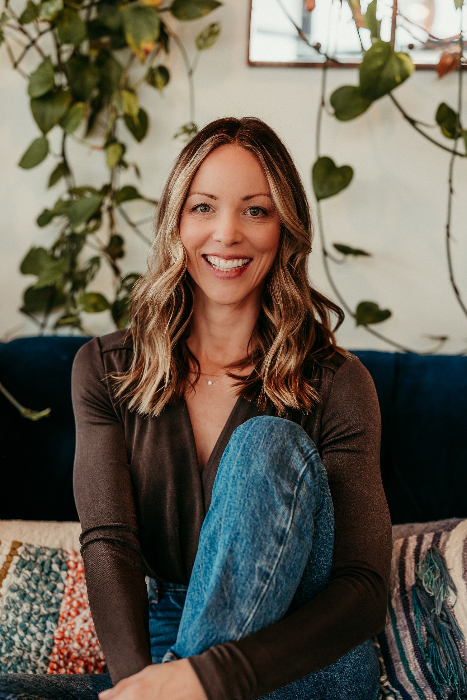 Smiling woman with wavy blonde hair sits on a couch in jeans and dark top, surrounded by lush green plants.