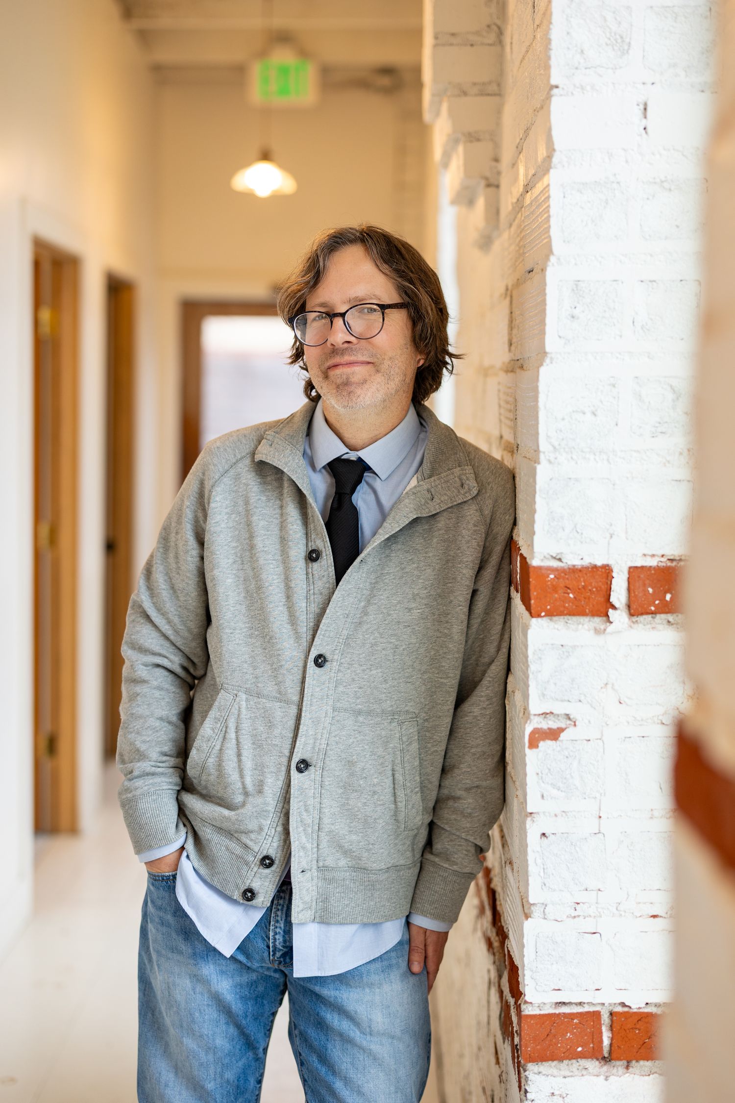 Middle-aged man with glasses and gray jacket leaning against a white brick wall in an office hallway.
