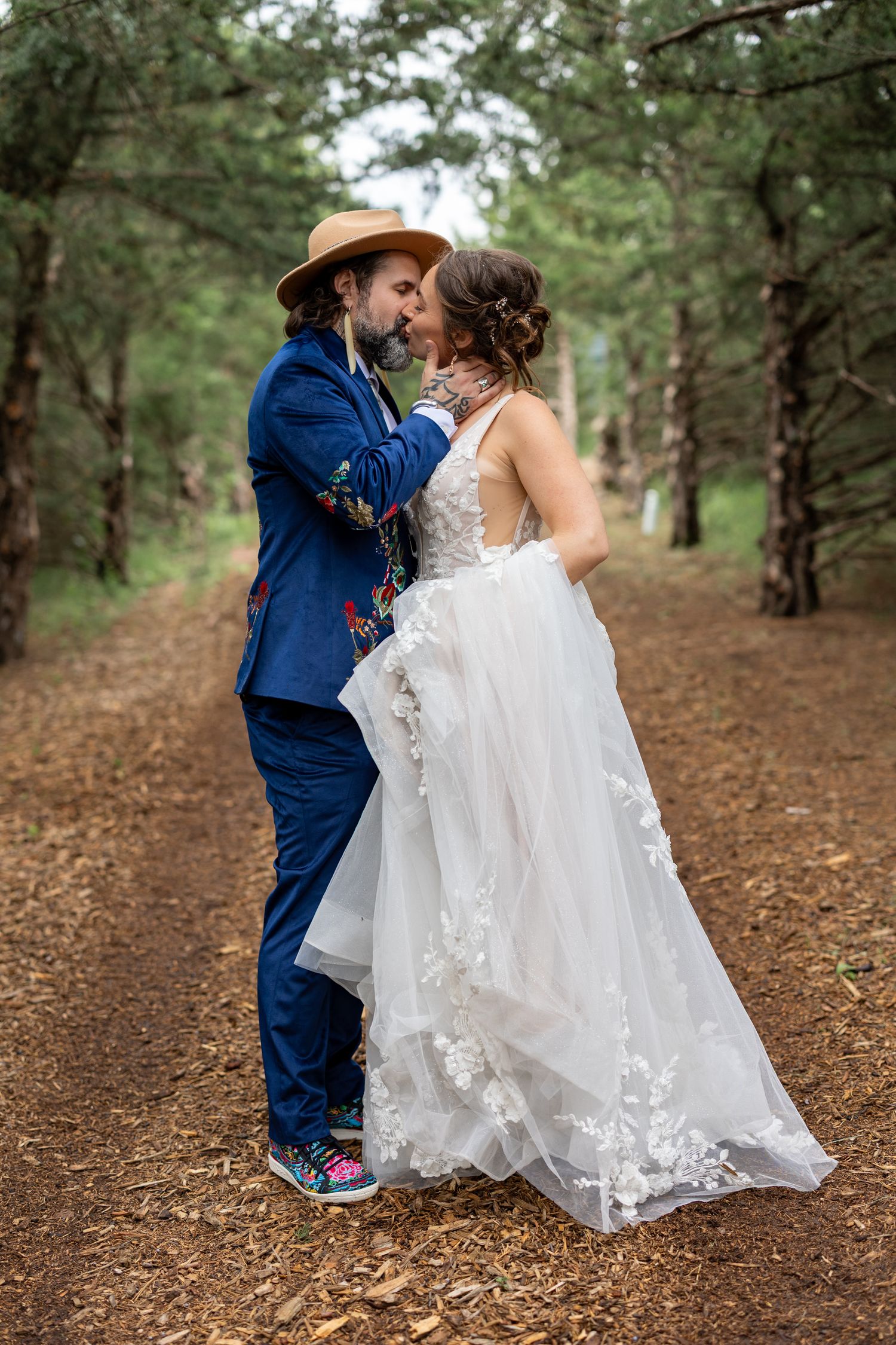 Bride and groom sharing a kiss in a pine forest, groom in blue suit and hat, bride in lace wedding gown.