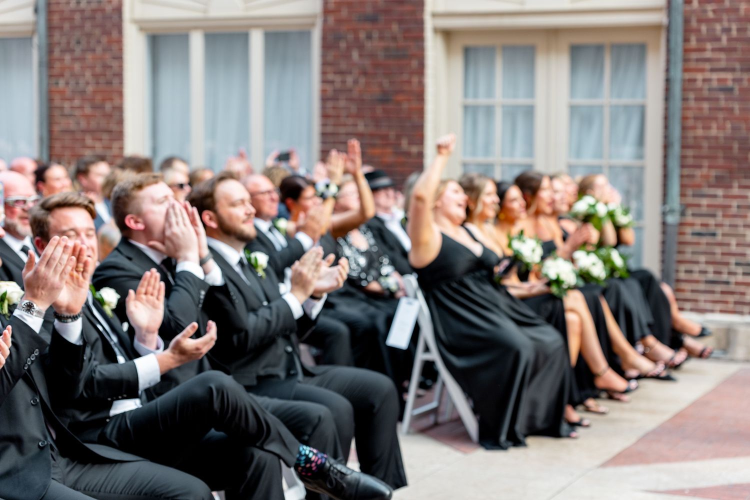 Wedding guests in black attire clapping and celebrating during an outdoor ceremony at a brick building venue.