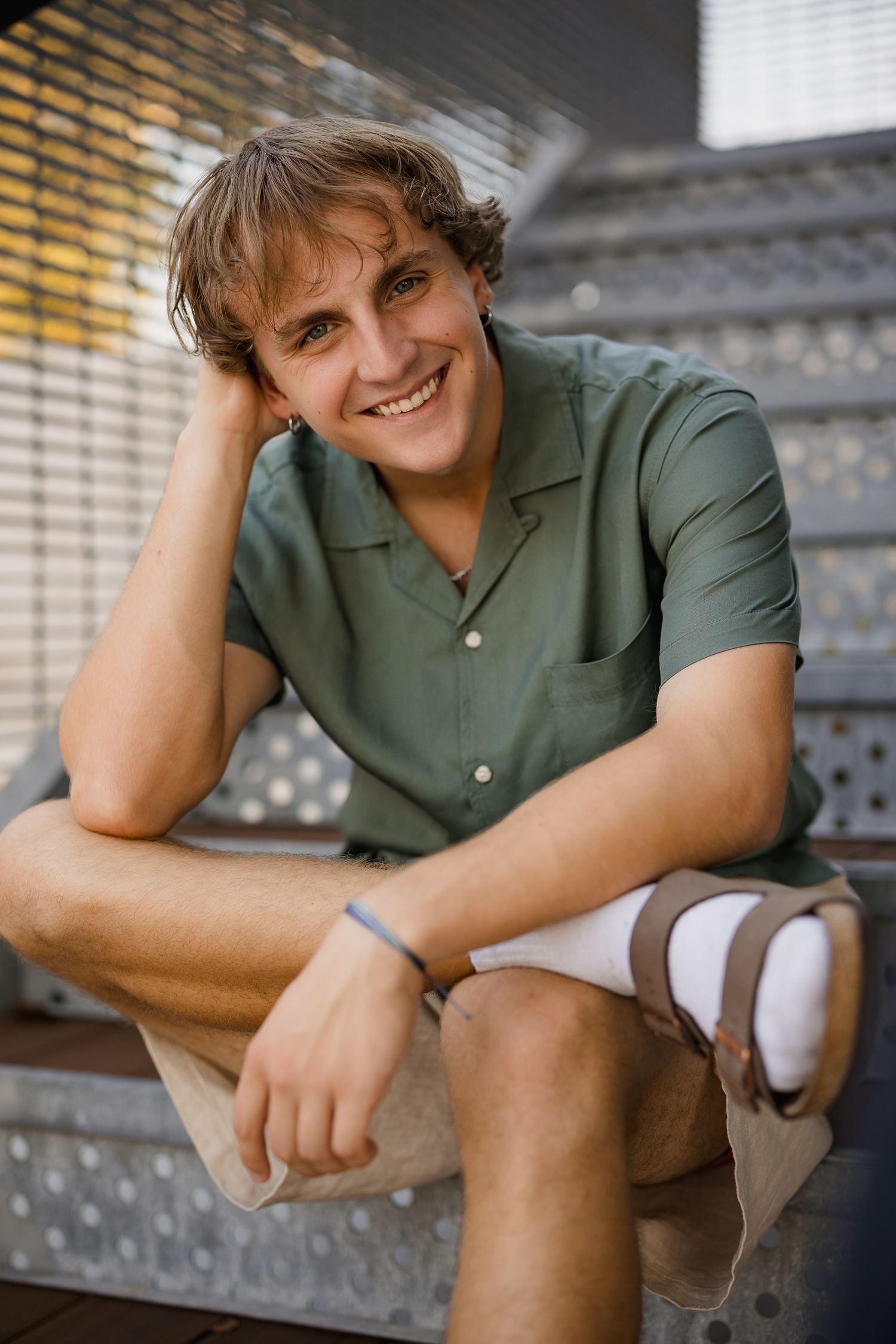 Smiling young man in green shirt and shorts sitting relaxed on steps, resting head on hand outdoors.