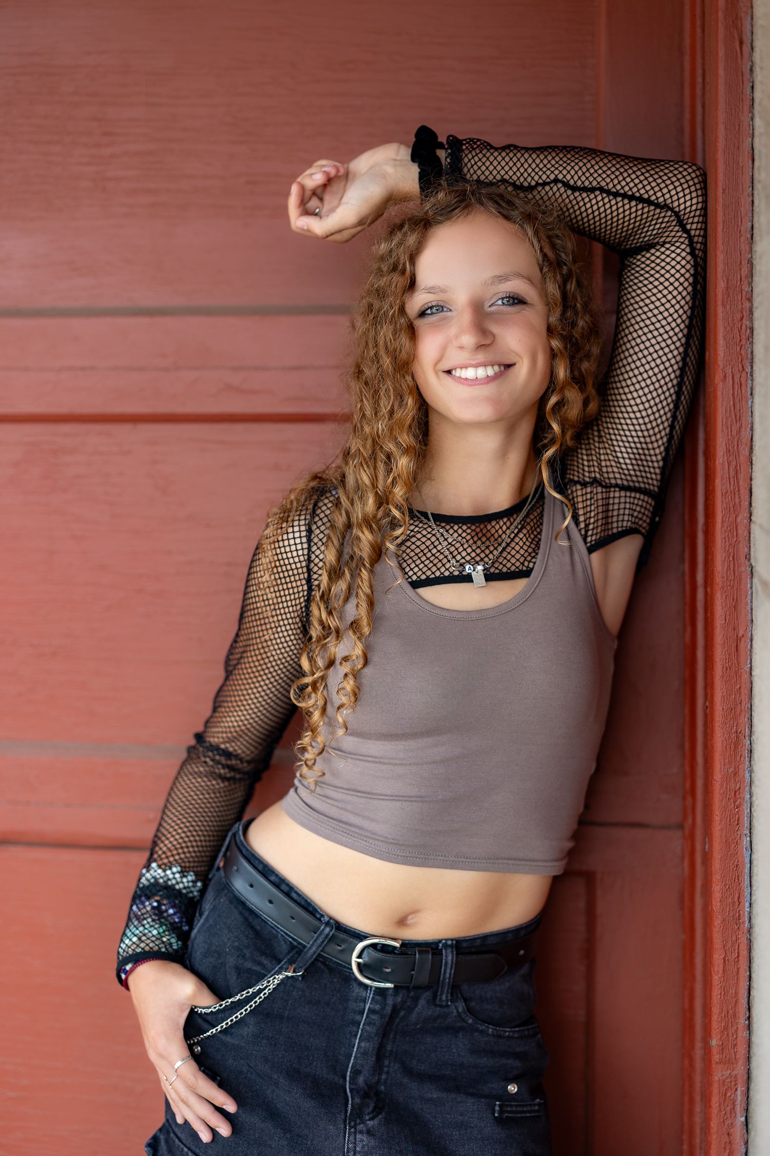 Smiling young woman with curly hair in crop top, fishnet sleeves, and jeans leaning against a red wall.