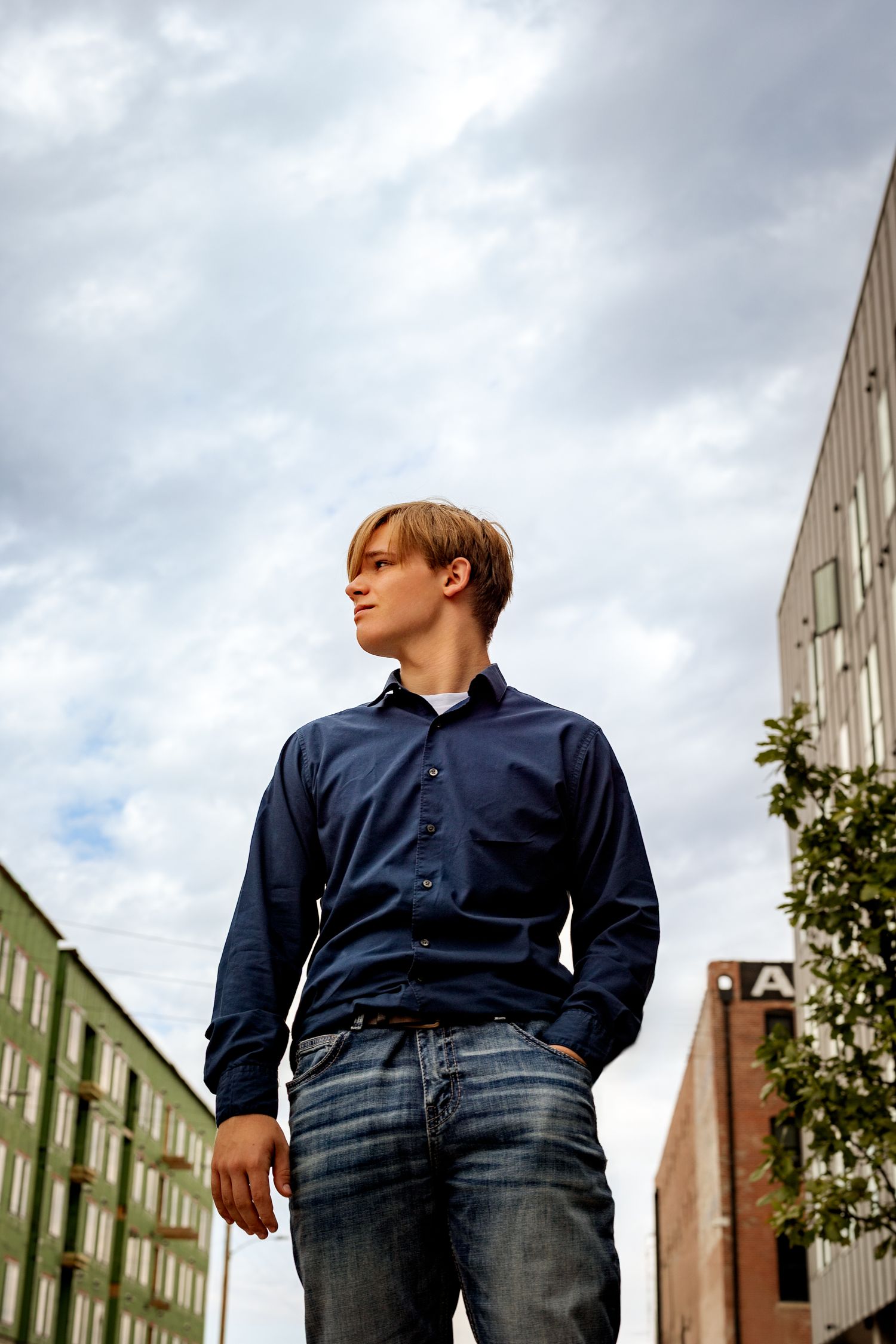 Young man in blue shirt and jeans standing outdoors against cloudy sky with urban buildings in background.