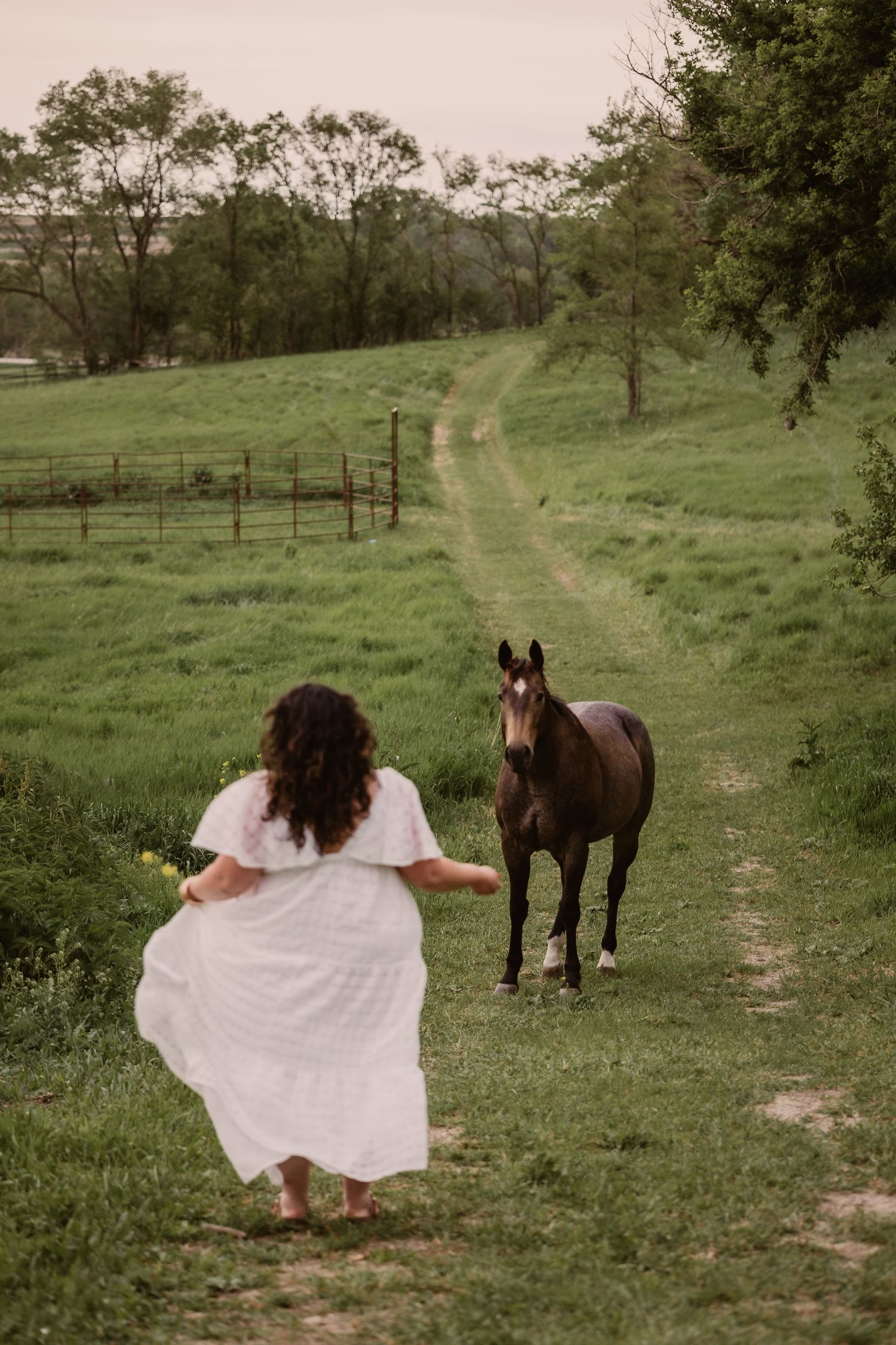A woman in a white dress runs barefoot toward a dark brown horse on a lush green farm path.