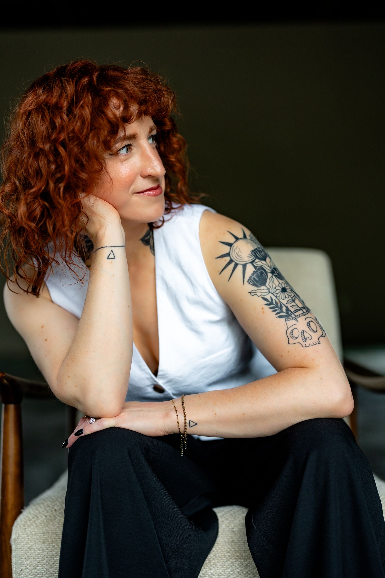 A redhead woman with curly hair and tattoos sits in a chair wearing a white sleeveless top and black pants.