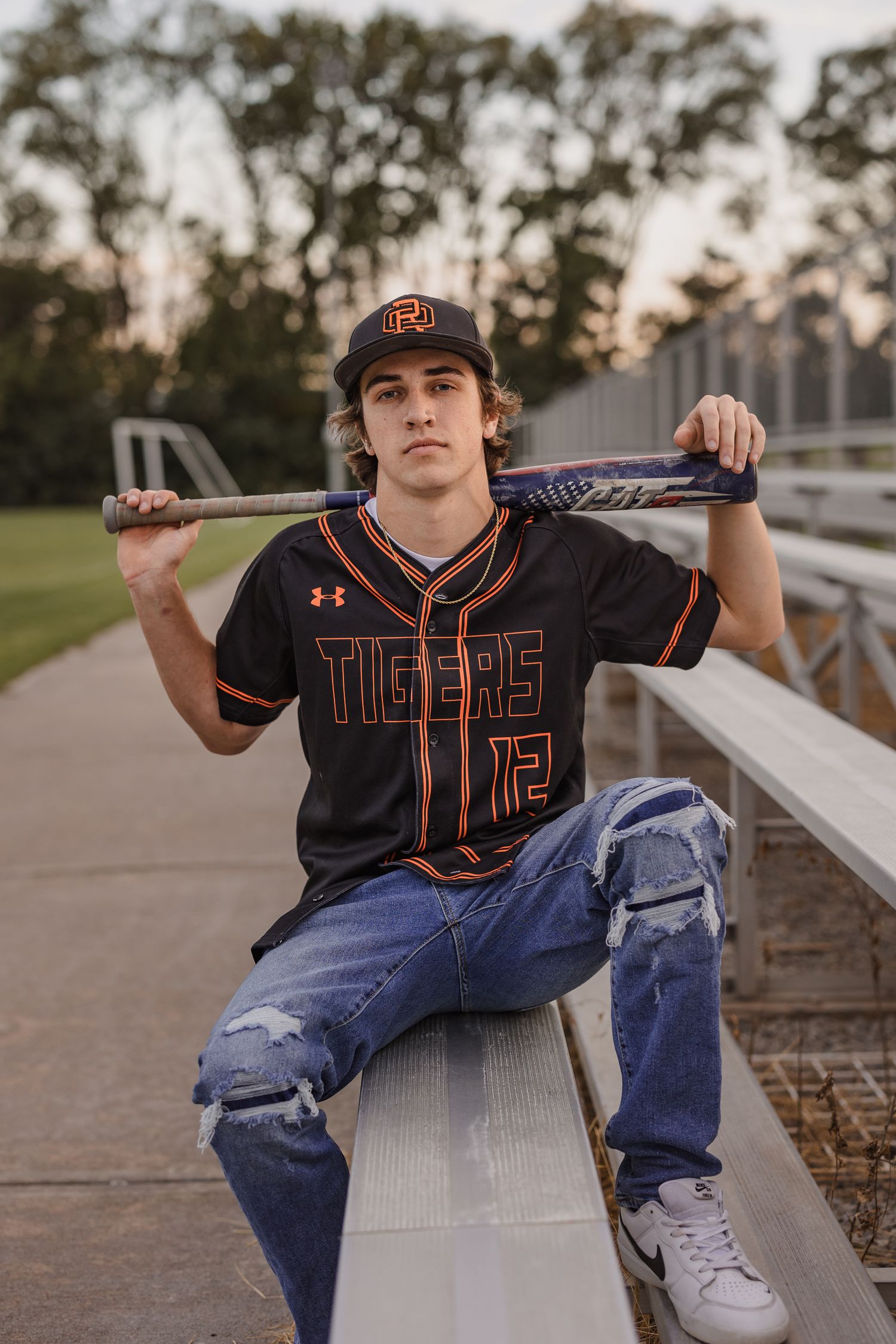 A teen in a Tigers baseball jersey holds a bat on his shoulders while sitting on bleachers in ripped jeans.