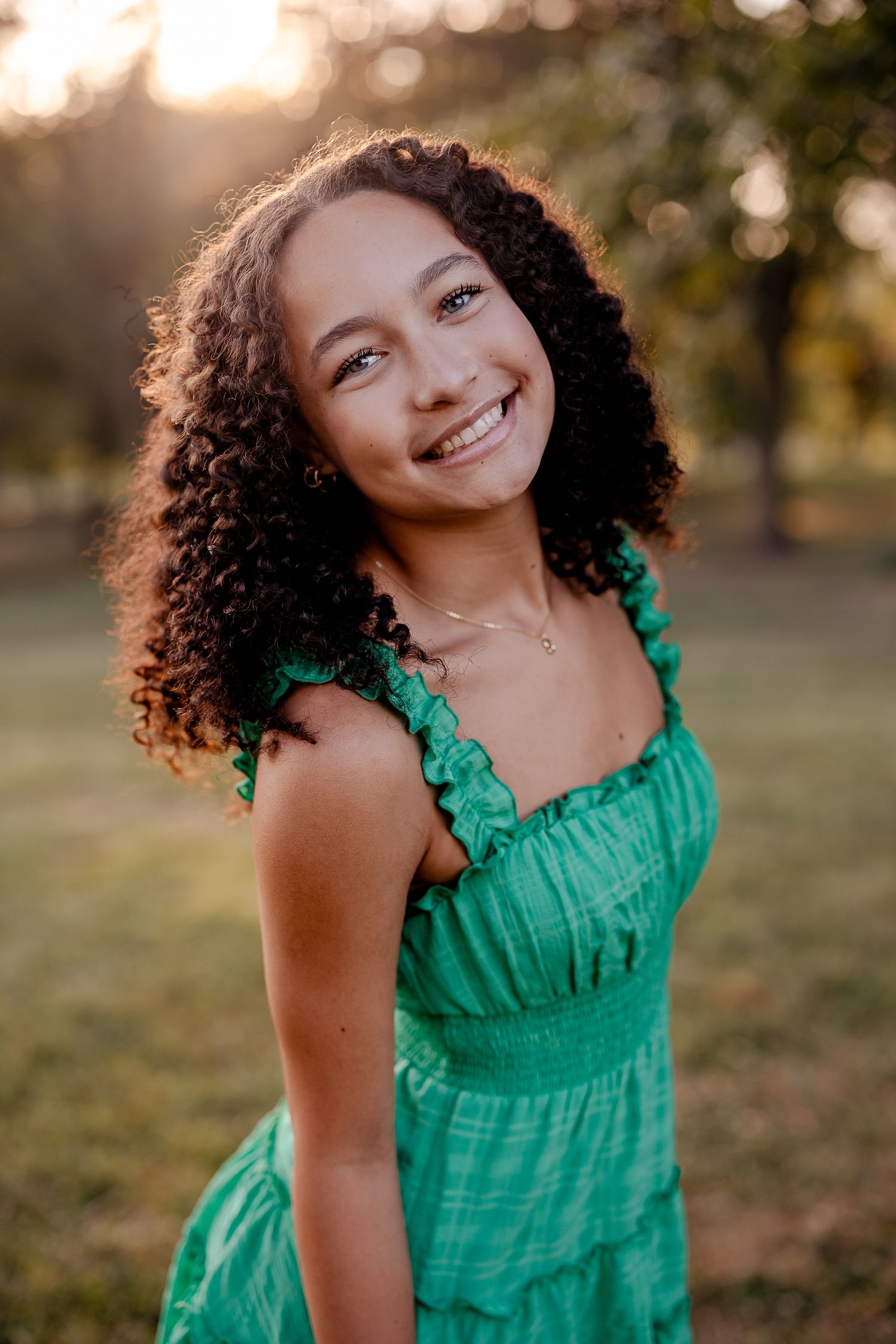 Smiling young woman with curly hair wearing a green ruffled dress outdoors in golden hour sunlight.
