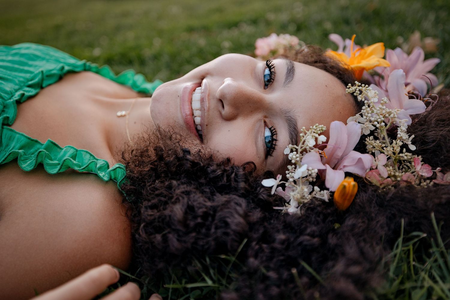 Young woman with curly hair lying on grass, adorned with pink and white flowers, smiling serenely in a green dress.