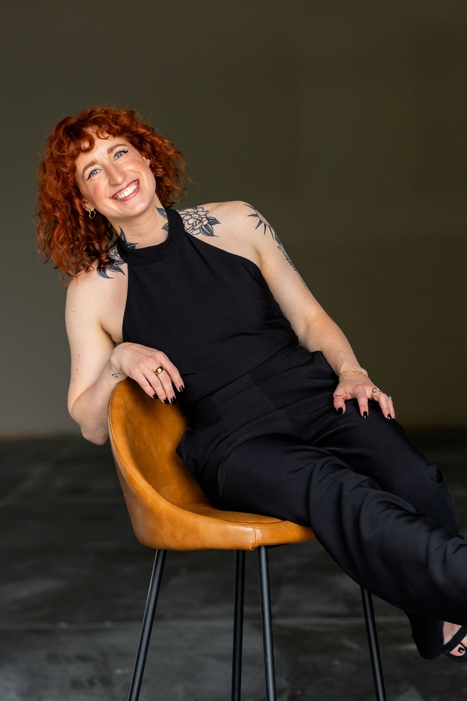 Smiling redhead woman with tattoos sits relaxed in leather chair wearing all-black outfit against dark background.