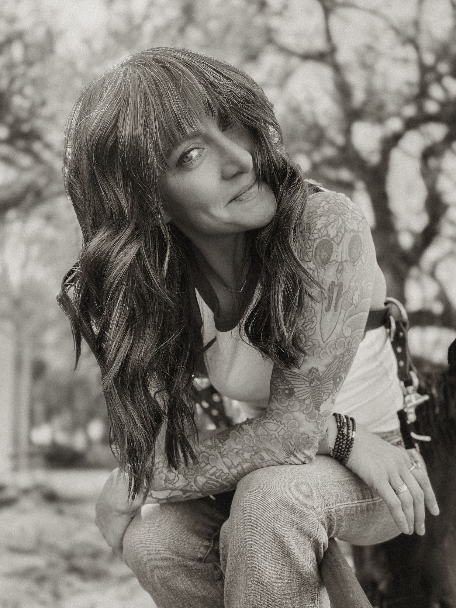 Black and white portrait of a tattooed woman with long wavy hair sitting outdoors, smiling softly.