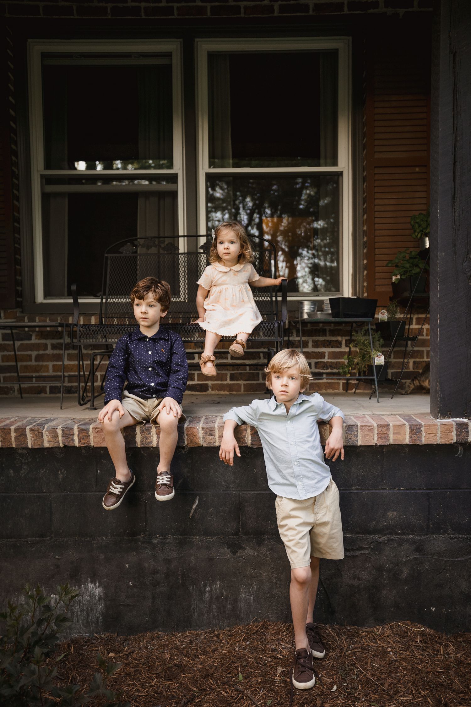 Three children pose on brick porch steps of a home, two boys and a toddler girl in a dress.