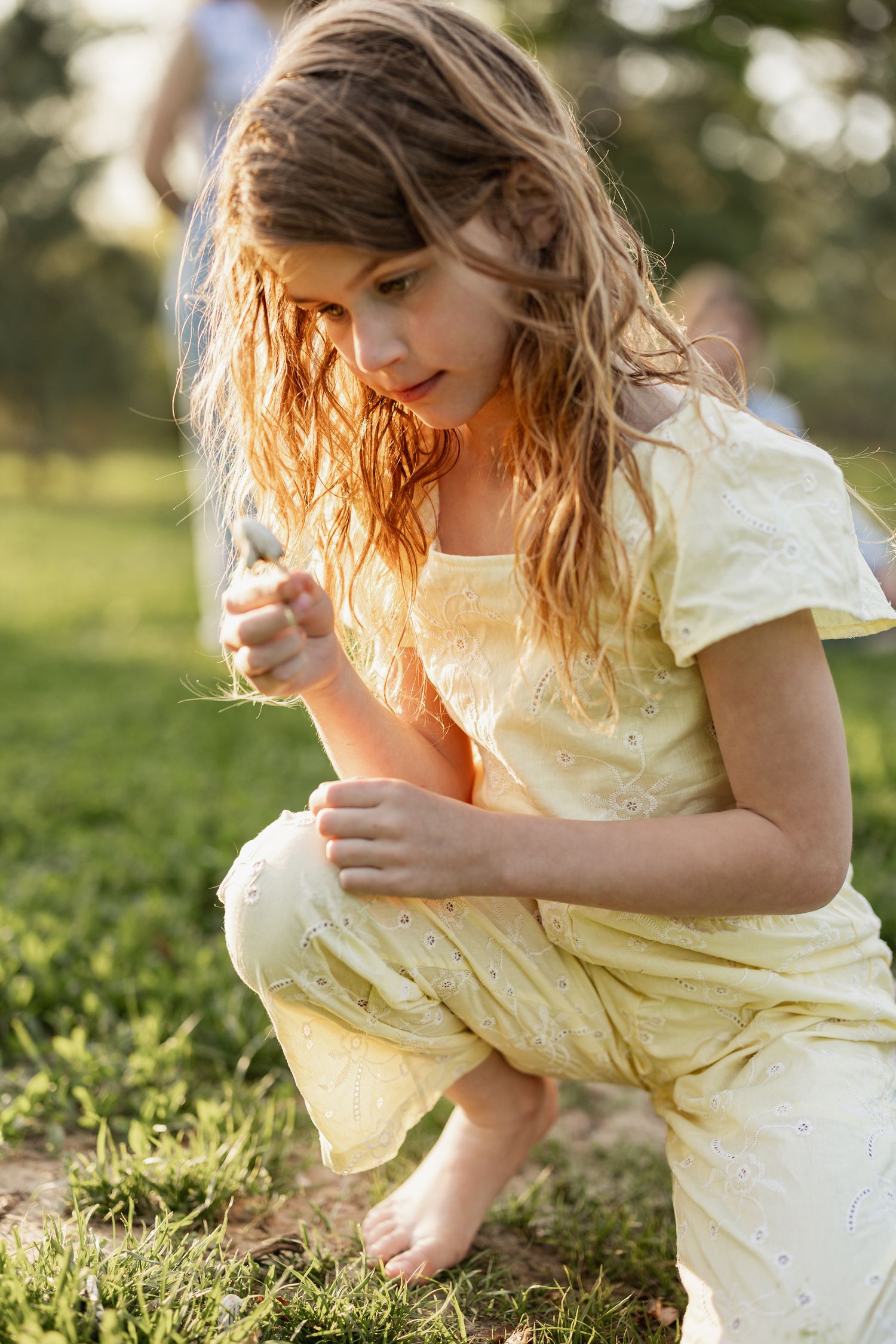 Young girl in yellow dress crouching barefoot on grass in golden sunlight, examining something in her hand.