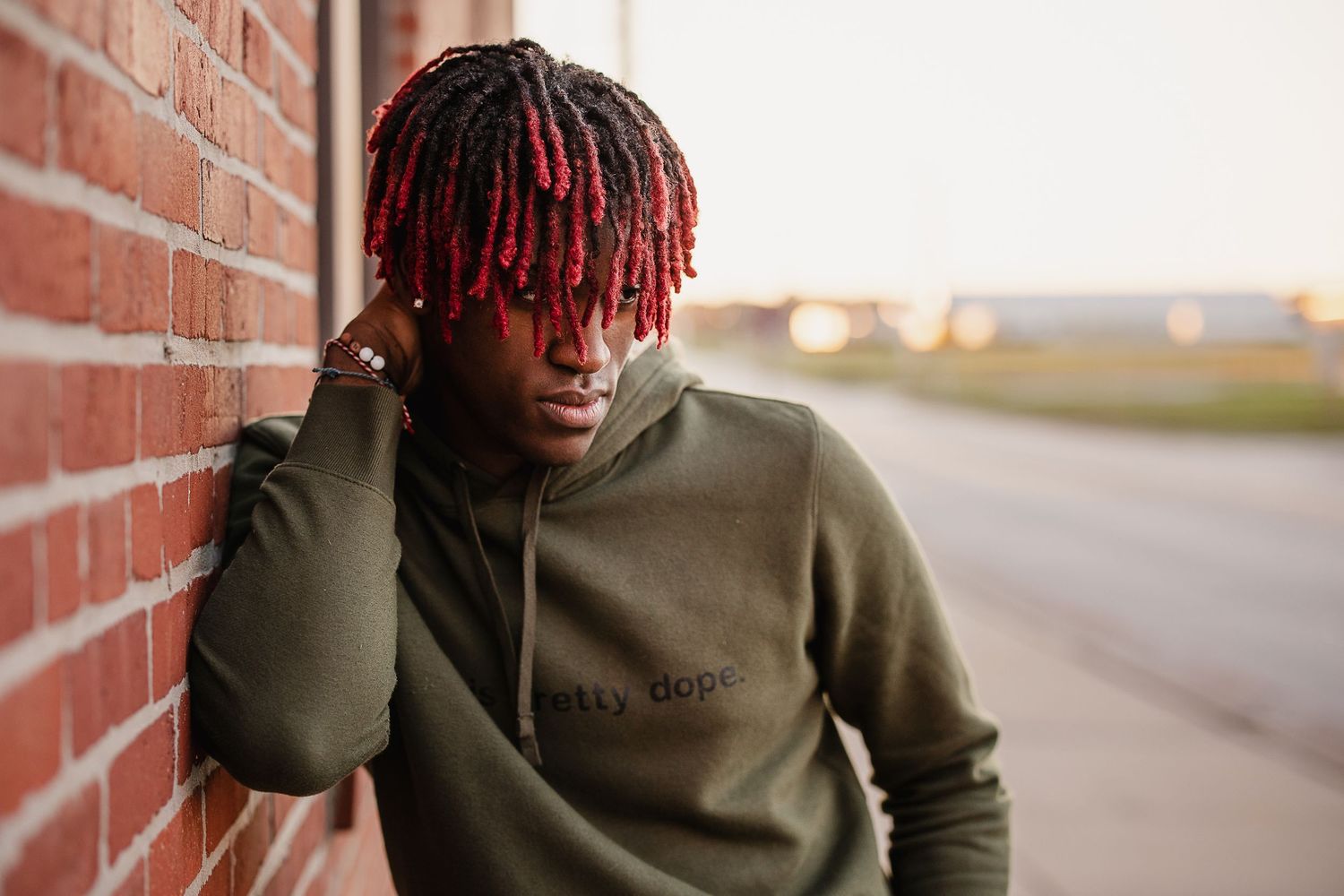 Young man with red dreadlocks wearing olive green hoodie leaning against brick wall outdoors.