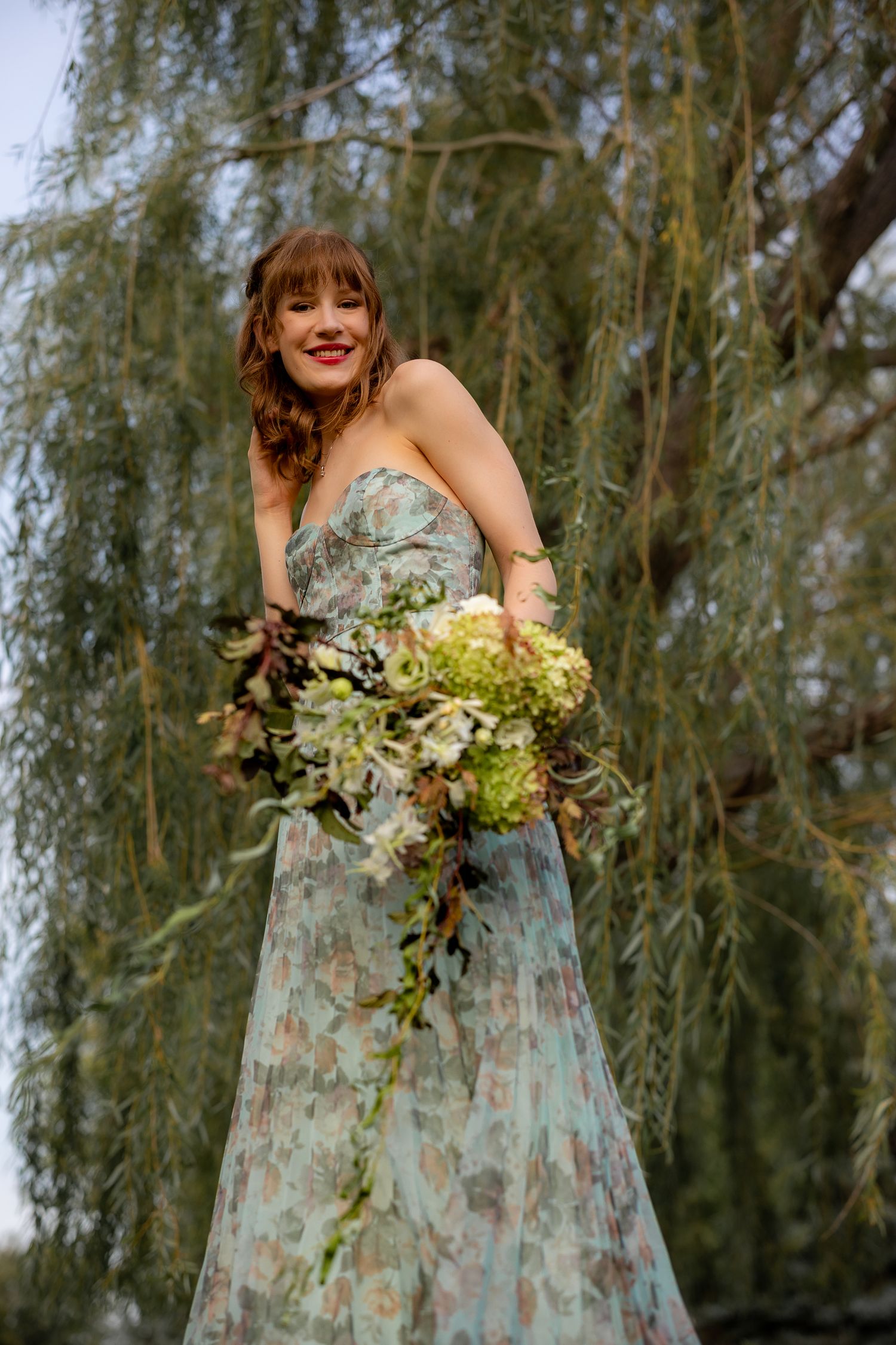 Smiling bride in floral strapless gown holds lush green bouquet under weeping willow tree outdoors.