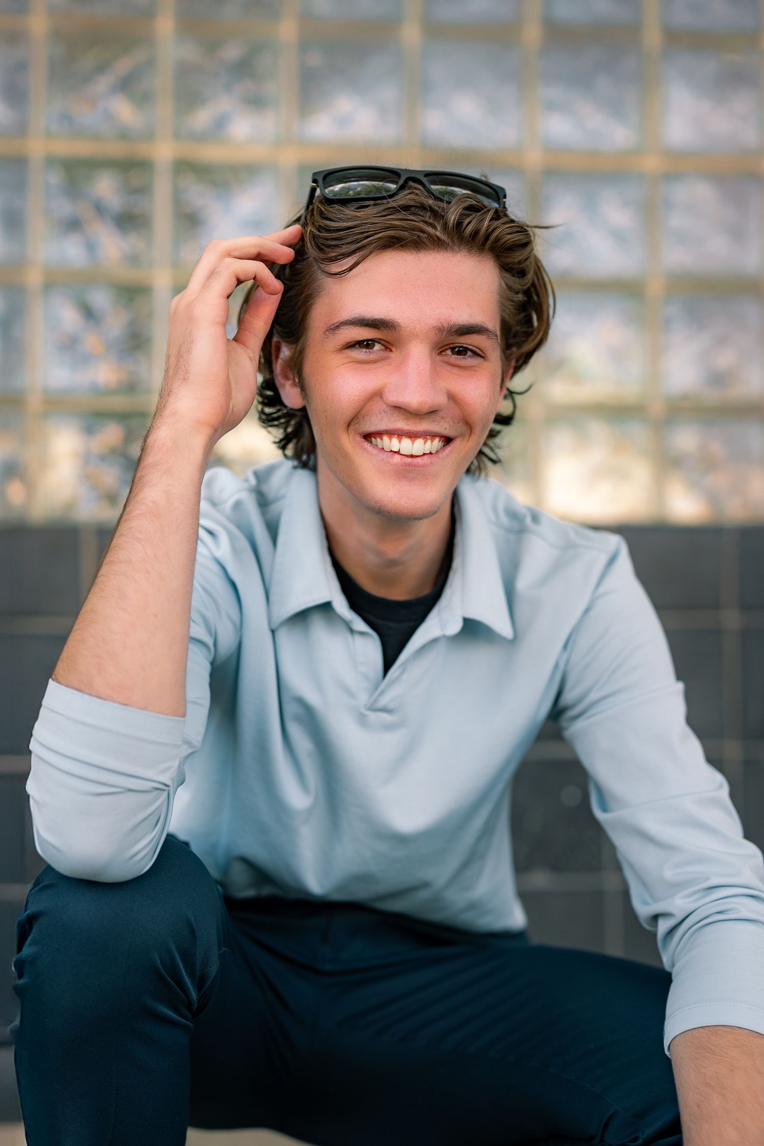 Smiling young man in light blue shirt with sunglasses on head, sitting outdoors against glass block wall.