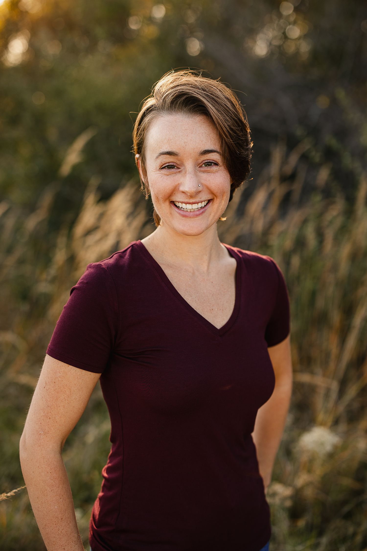 Smiling woman in maroon v-neck shirt posing outdoors in golden hour light with tall grasses in background.