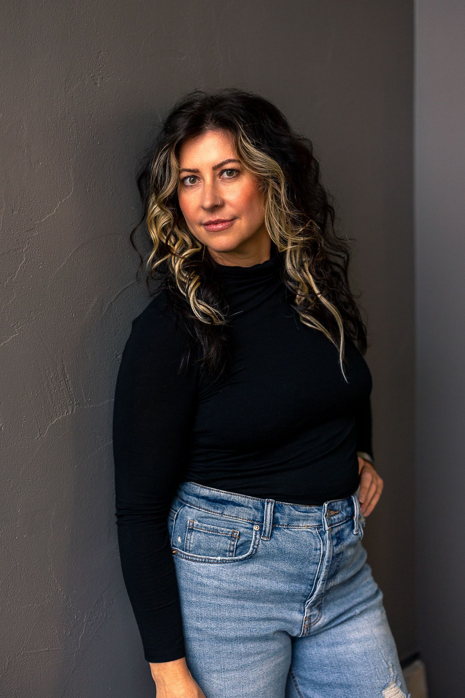 Woman with dark wavy highlighted hair posing in black turtleneck and light ripped jeans against gray wall.
