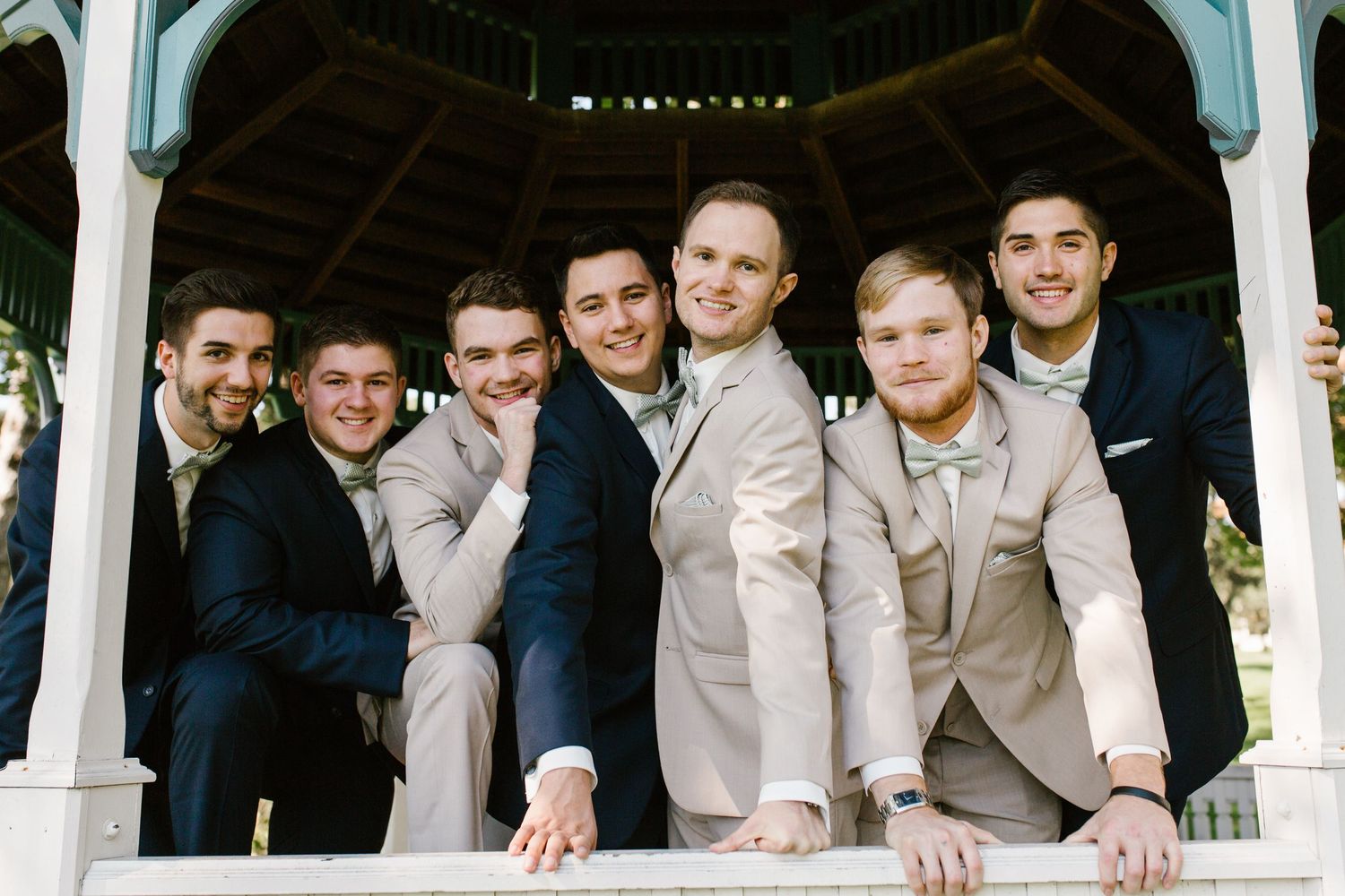 Two grooms and their groomsmen in navy and tan suits posing together under a white gazebo at an outdoor wedding celebration.