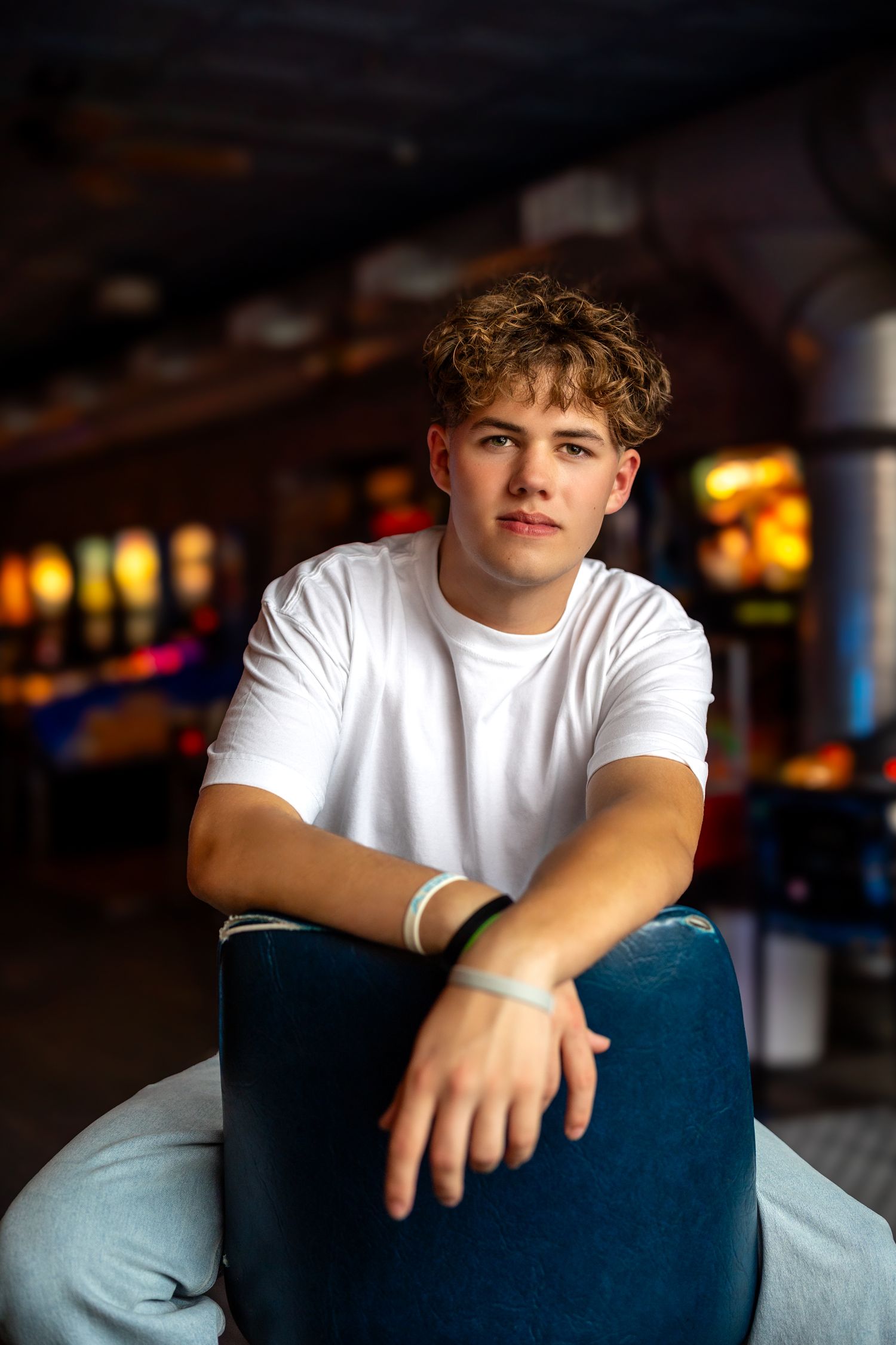 Young man with curly hair in white t-shirt sits relaxed in blue chair, colorful bokeh background.