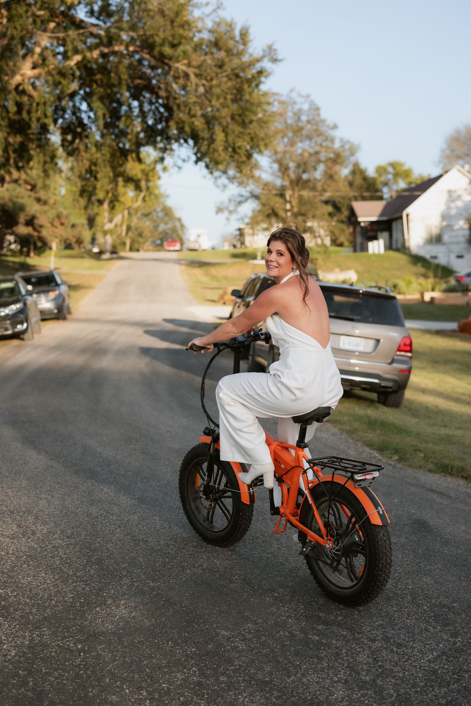 Woman in white romper wedding outfit riding an orange electric fat tire bike on a suburban street, smiling over her shoulder.