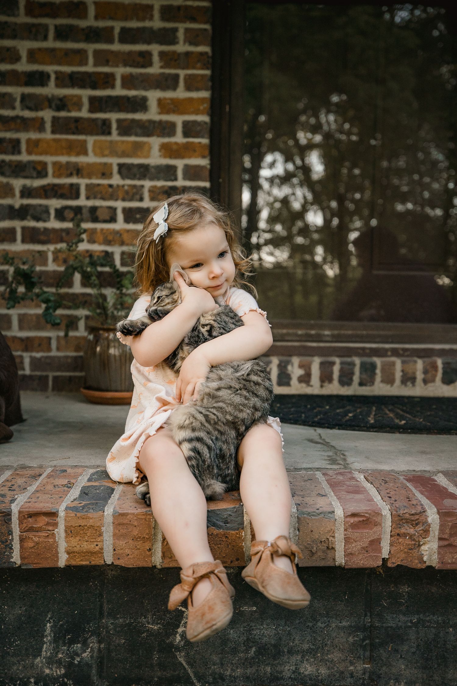 Young girl in pink dress hugging a tabby cat on brick steps outside a home.