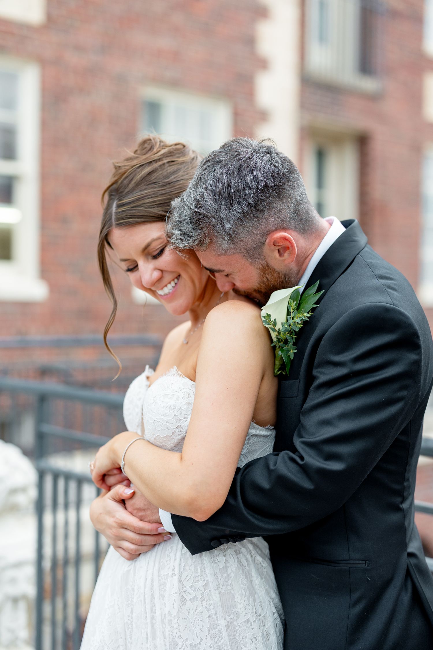A smiling bride in a strapless lace gown embraces her groom in a black tuxedo with boutonniere outside a brick building.