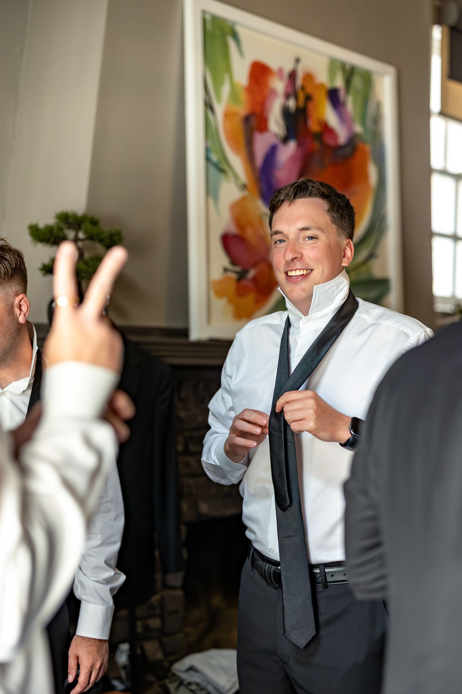 Groom smiling while adjusting his tie, surrounded by groomsmen in a elegant room with colorful floral artwork.
