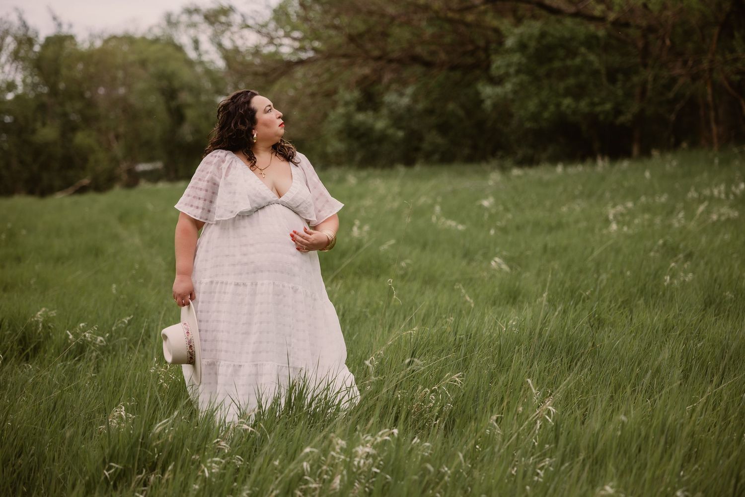 Woman in white lace dress walks through lush green meadow, holding a hat at her side, gazing upward peacefully.