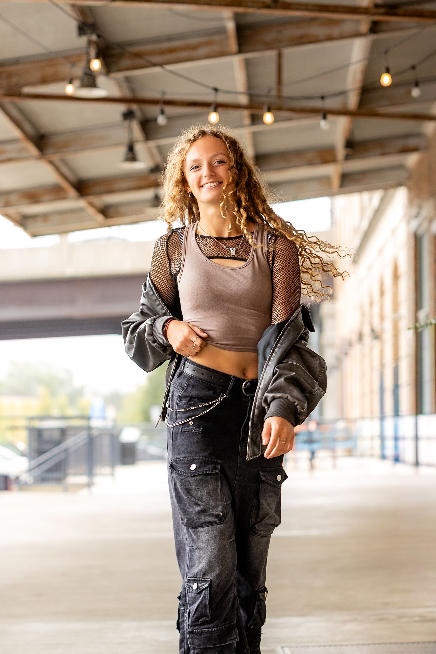 Smiling young woman with curly hair in crop top, mesh shirt, cargo jeans, and jacket walking under covered urban walkway.