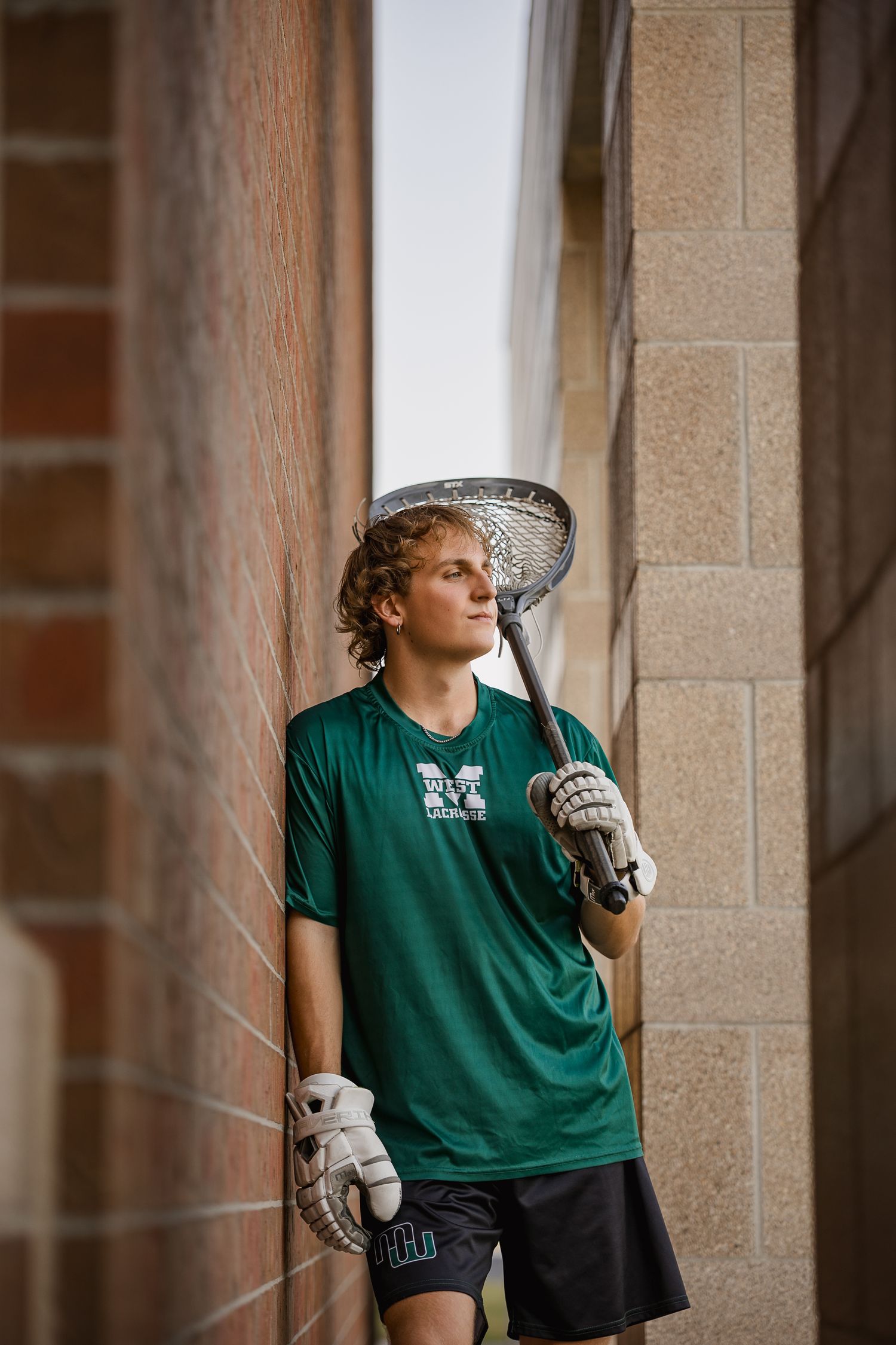 Young lacrosse player in green jersey holding stick and gloves, leaning against brick wall outdoors.