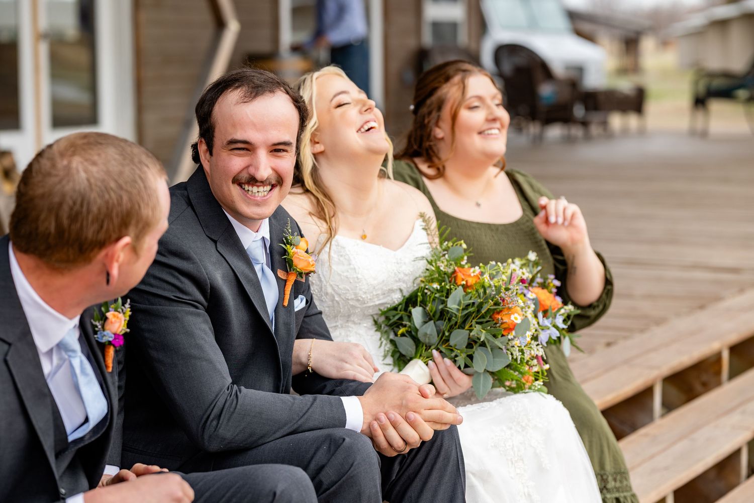 Bride and groom laughing joyfully with guests on wooden steps, bride holding lush green and orange bouquet.