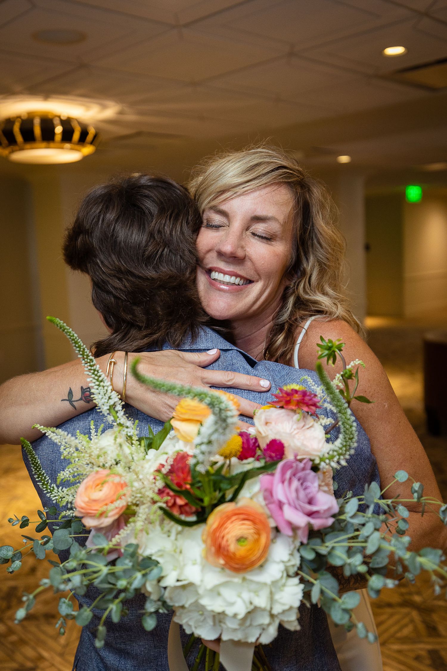 Smiling blonde woman hugging someone while holding colorful bridal bouquet at indoor venue.