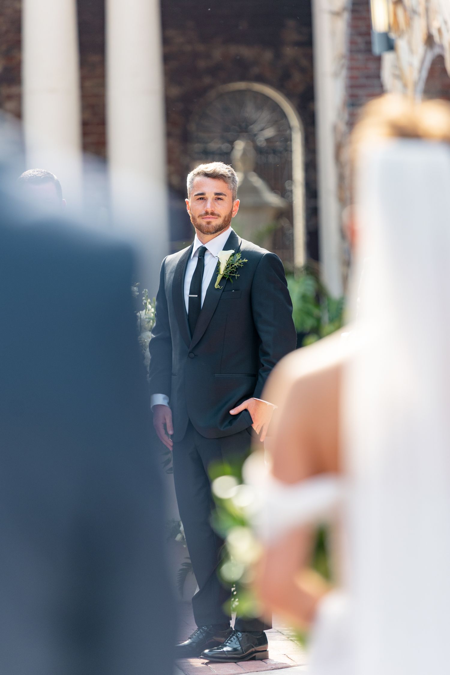 Groom in dark suit with boutonniere stands at altar waiting as bride approaches during outdoor wedding ceremony.