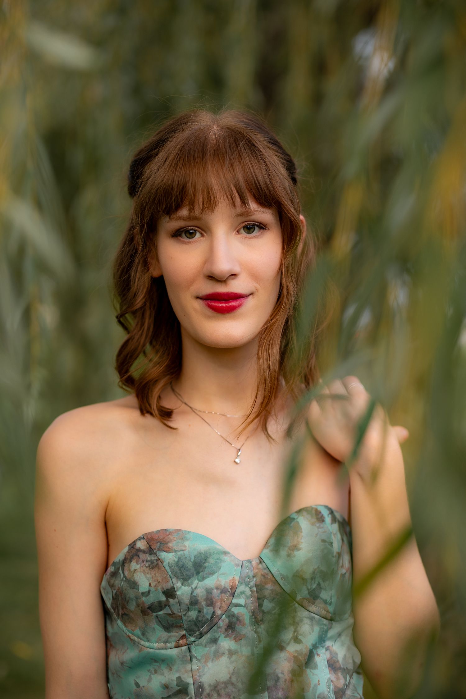 Young woman with red lips in floral strapless dress poses among green willow branches outdoors.