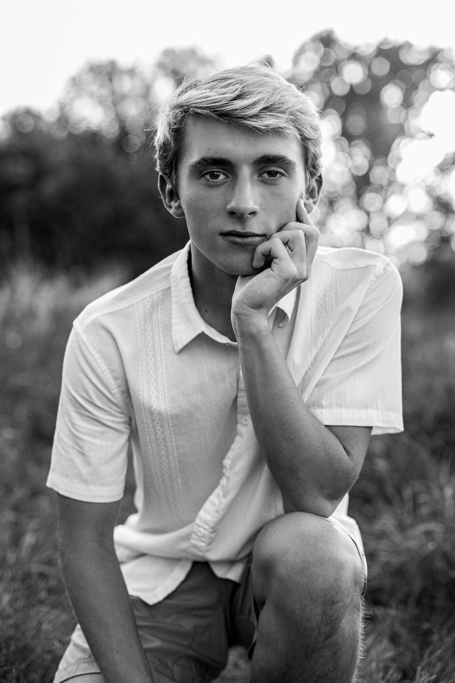 Black and white portrait of a young blonde teen boy in a white shirt sitting outdoors in a field.