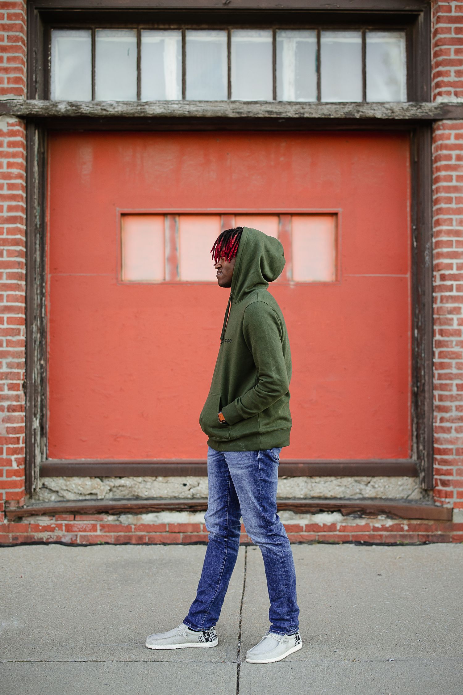 Young Black man in green hoodie, blue jeans, and white sneakers walking on urban sidewalk near red brick building.