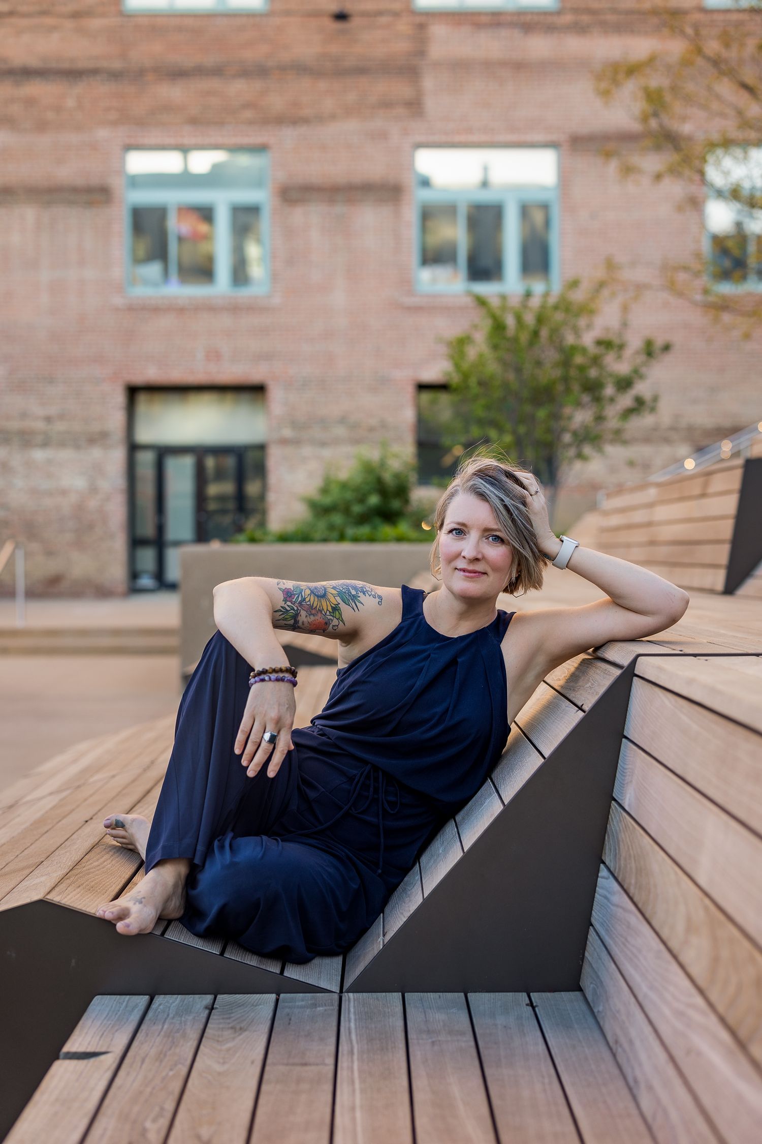 Woman in navy jumpsuit with tattoos relaxes on modern wooden outdoor bench with brick building backdrop.