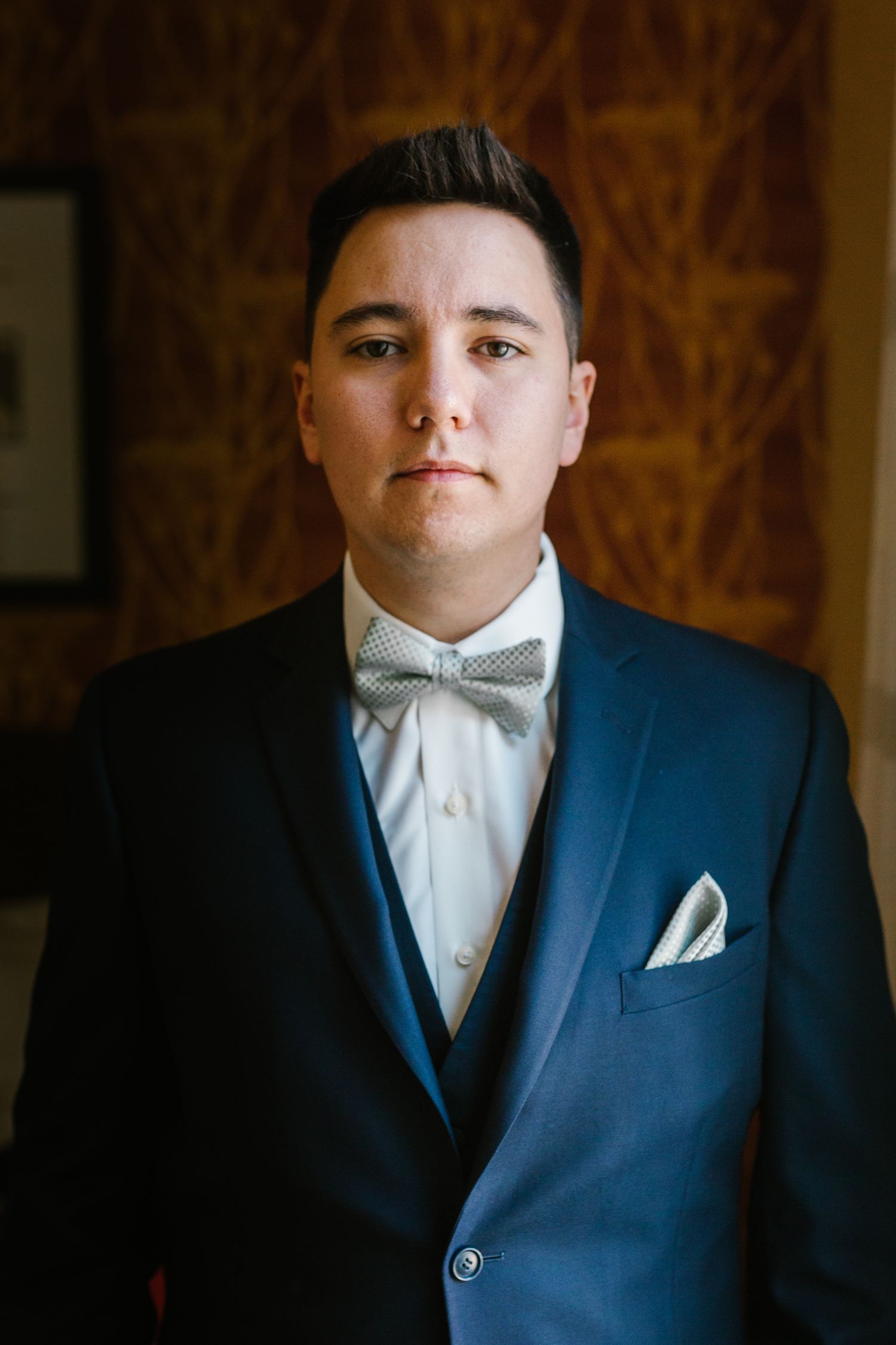 Young man in blue suit with gray bow tie and pocket square posing formally indoors.